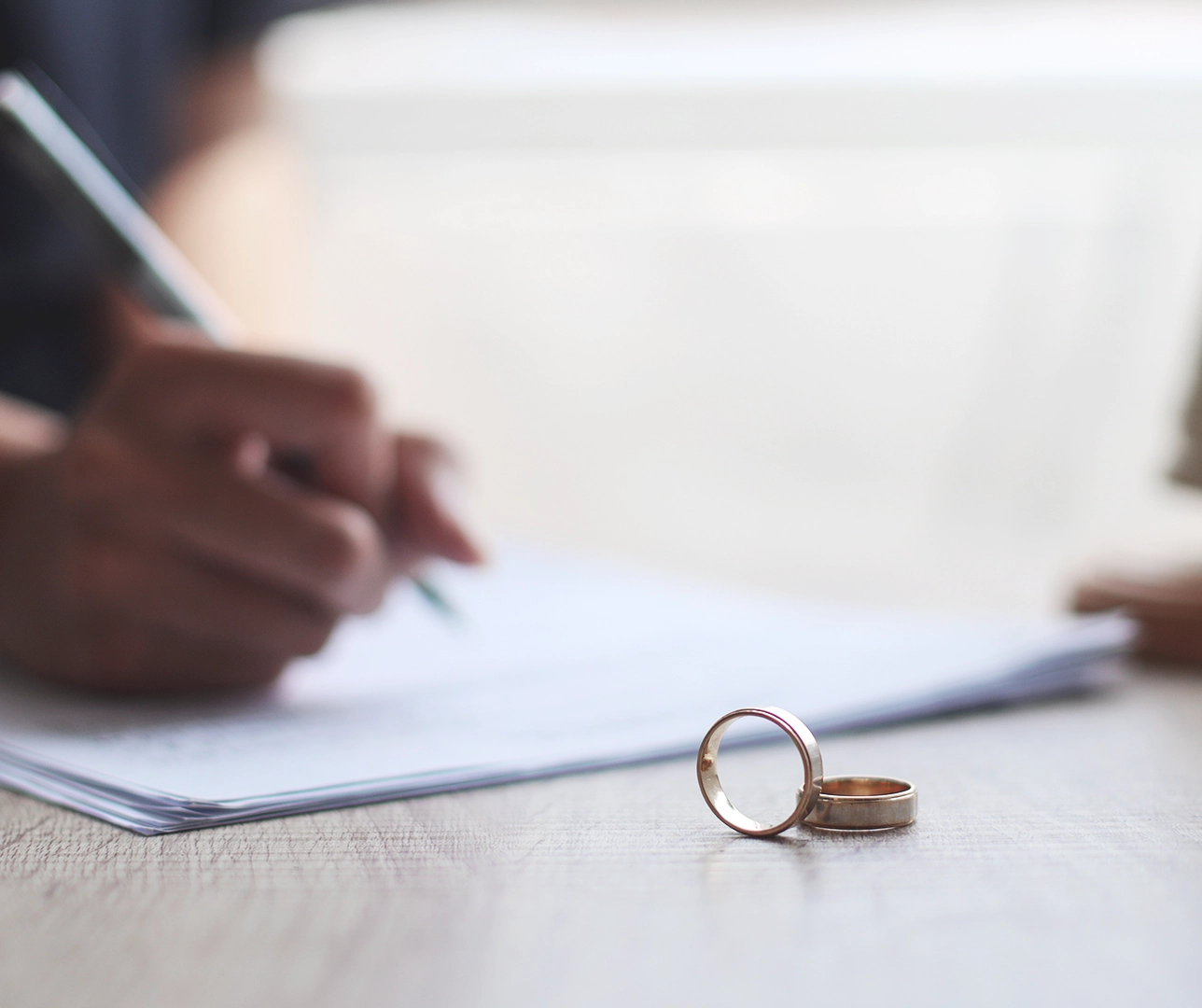 Unos anillos en primer plano y una persona firmando el divorcio de fondo.